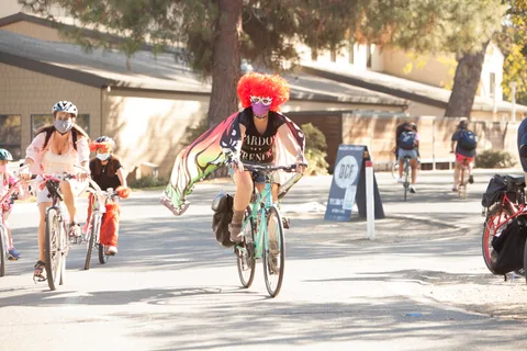 A rider in an orange wig and butterfly wings pedals hard with the cape flying behind.