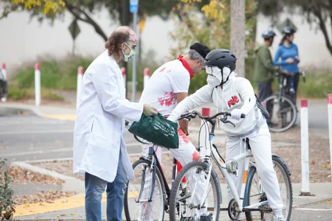Two riders dressed as bloodied doctors in white coats stand together with their bikes.