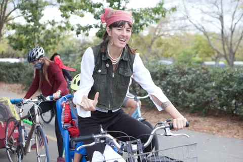 A masked rider in a pirate vest gives a thumbs up while holding her bike.