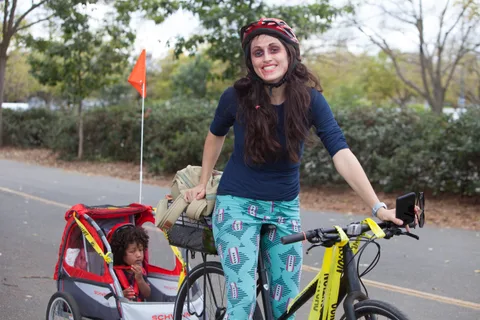 A woman stands by her bike with a child in a small trailer flying an orange flag.