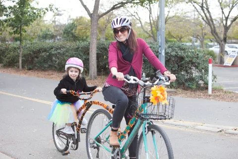 A mother and daughter in a tutu smile while riding a tandem bike on a park path.