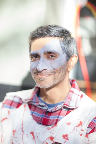 A man in a bloodstained plaid shirt smiles with fake wound makeup.