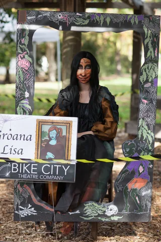 A woman in dramatic dark makeup poses under a decorated Halloween archway.