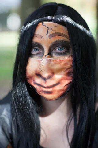 A woman with striking blue eyes wears cracked porcelain doll makeup and a matching mask.