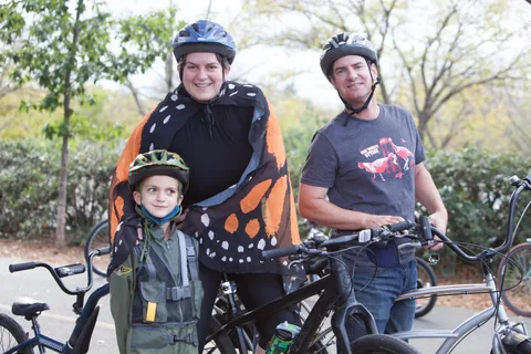 Two men in costume stand with a child and their bikes, one wearing an orange cape.