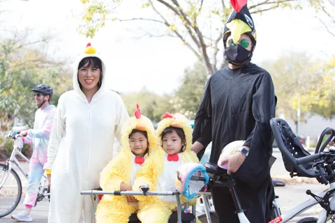 A rider in a white bunny costume waits on a cargo bike with kids in yellow chick costumes.