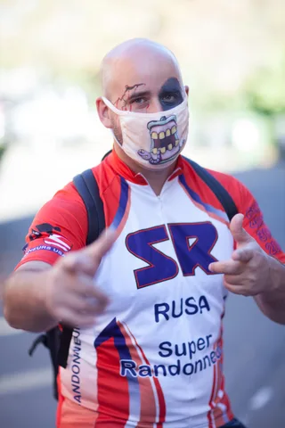A man in a cycling jersey with wound makeup and a monster-mouth mask points at the camera.