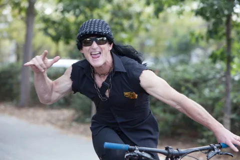 A woman in a black costume strikes a playful rock-and-roll pose on her bike.