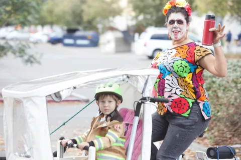 A woman in a colorful harlequin costume rides a cargo bike with a child in a hooded costume.