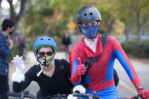 A man in a full Spider-Man costume gives a thumbs up beside a friend in a skeleton hoodie.