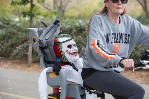 A woman rides a cargo bike with a masked figure strapped into the child seat.