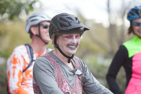 A rider in zombie face paint laughs while other costumed cyclists wait behind her.