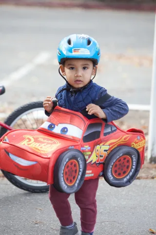A toddler in a blue helmet wears a full Lightning McQueen race car costume.