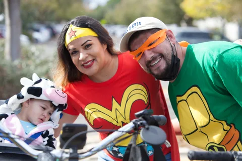 A family in Wonder Woman and superhero costumes leans in together with a baby in a cow costume.