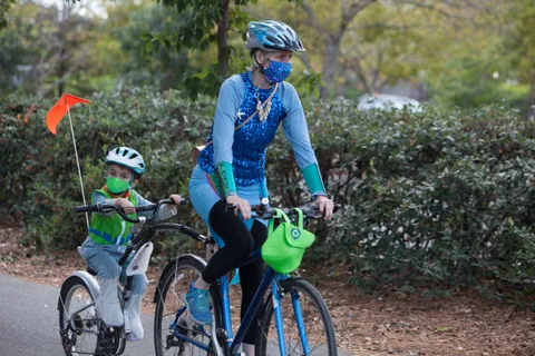 A child rides ahead with a tall orange safety flag while a masked adult follows.