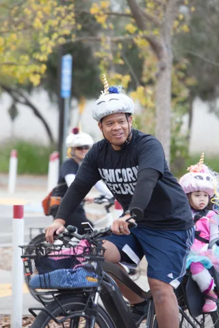 A rider in a unicorn party hat waits on a bike path with a child behind him.