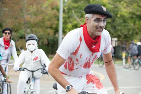 A man in a bloodstained apron and red bandana stands with masked skeleton kids behind him.