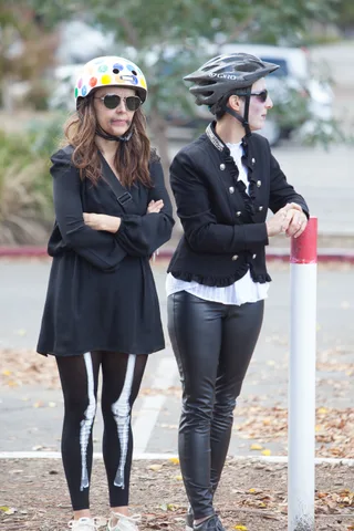 Two women in black skirts with painted skeleton legs walk away from the camera.