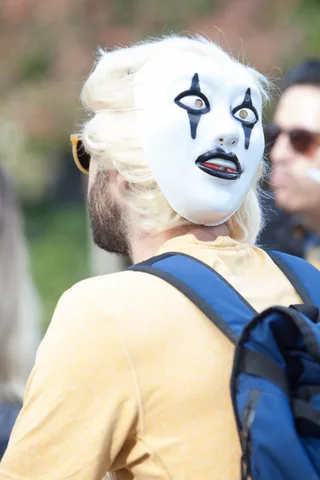 A man wears an eerie white mask on the back of his head, facing backward.