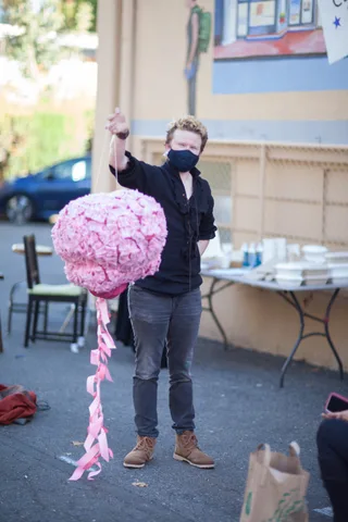A man in a dark mask raises a large pink pom-pom decoration overhead.