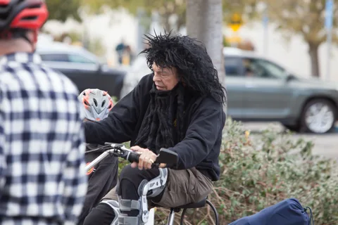 A rider with a long dark wig and dramatic makeup poses on a bike near a parked car.