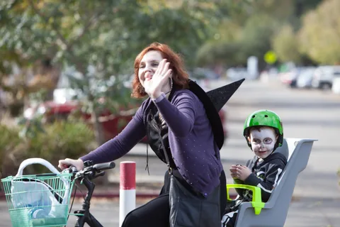 A woman in a witch costume waves while a child in a green helmet rides in the bike seat behind her.