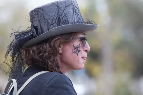 A woman in a lace top hat with a spiderweb face tattoo looks off to the side.