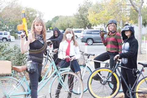 A group in dark punk-styled costumes and masks stands together with their bikes.