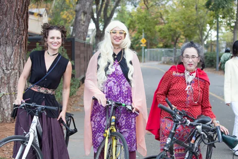 Three women in flowing costumes pose together with their bikes on a tree-lined street.