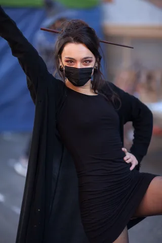 A woman in black raises her arm dramatically, face partly covered by a black mask.