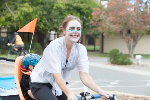 A woman in white skeleton face paint smiles while riding with a small orange flag on her bike.