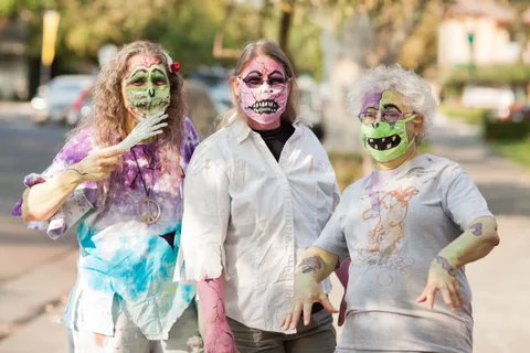Three costumed zombies in painted masks pose together on a sunlit sidewalk, one pointing a skeleton hand.