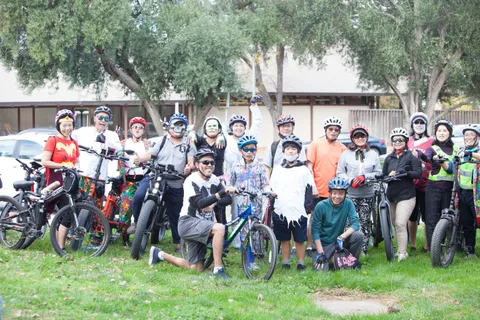 A large group of costumed riders and families gathers with bikes on the grass.