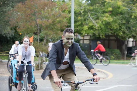 A man in a blazer costume adjusts his bike as another rider passes in a blur.
