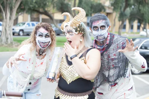 Three zombie-costumed friends react with excitement as one holds a Best Costume trophy.