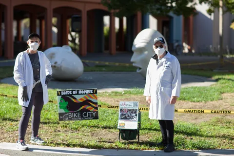 Two masked volunteers in doctor costumes stand near Bride of Zombie and NorCal Trykers donation signs.