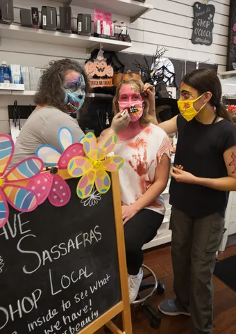 A volunteer paints flowers on a woman's face at a Halloween face-painting booth.
