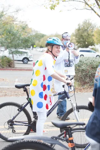 A rider in a colorful polka dot costume stands with her bike on the path.