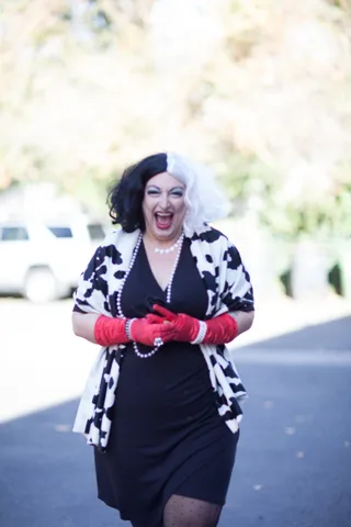 A woman in a black and white cow-print jacket and pearls laughs with pink gloved hands raised.