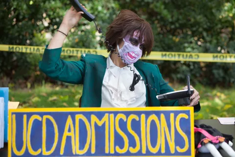 A zombie-costumed office worker answers phones behind a hand-painted UCD Admissions sign.