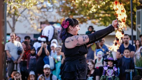 A fire performer with tattooed arms swings a flaming prop above a watching crowd at golden hour.