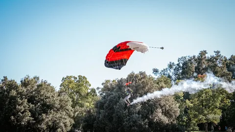 A skydiver glides in under a striking red and black parachute with a long smoke trail against a clear blue sky.