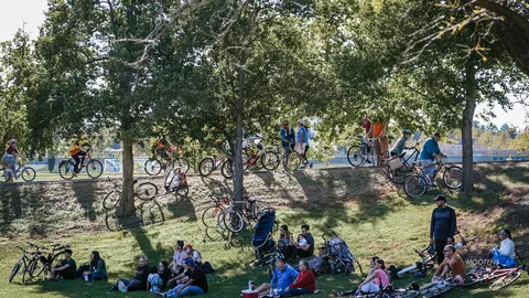 Families rest on a grassy park slope while riders pass by on the path above.