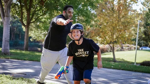 A dad in zombie makeup and his laughing son in an Ink Monkey shirt run together with a toy blaster.