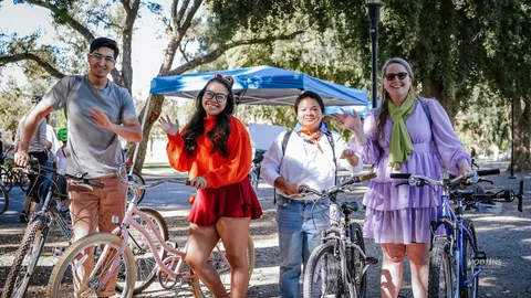 Four friends in matching cartoon-inspired costumes wave together with their bikes under golden park light.