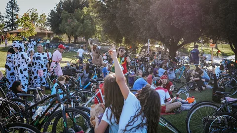 A costumed crowd with parked bikes gathers under the trees.