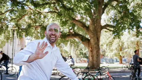 A man with green face paint grins and waves under a large shade tree.