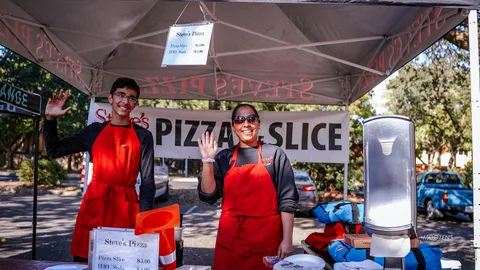 Two Steve's Pizza staff wave from behind their booth counter.