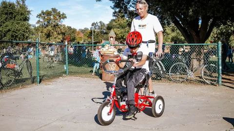 A boy in a helmet sits on a red adaptive trike beside a volunteer.
