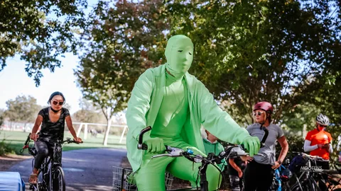 A rider in a full green bodysuit and mask pedals through the park past other cyclists.